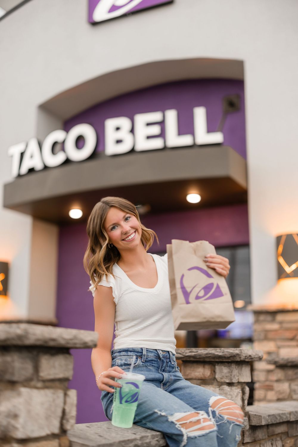 A customer sits with a takeout bag outside a Taco Bell restaurant storefront with purple signage at dusk.
