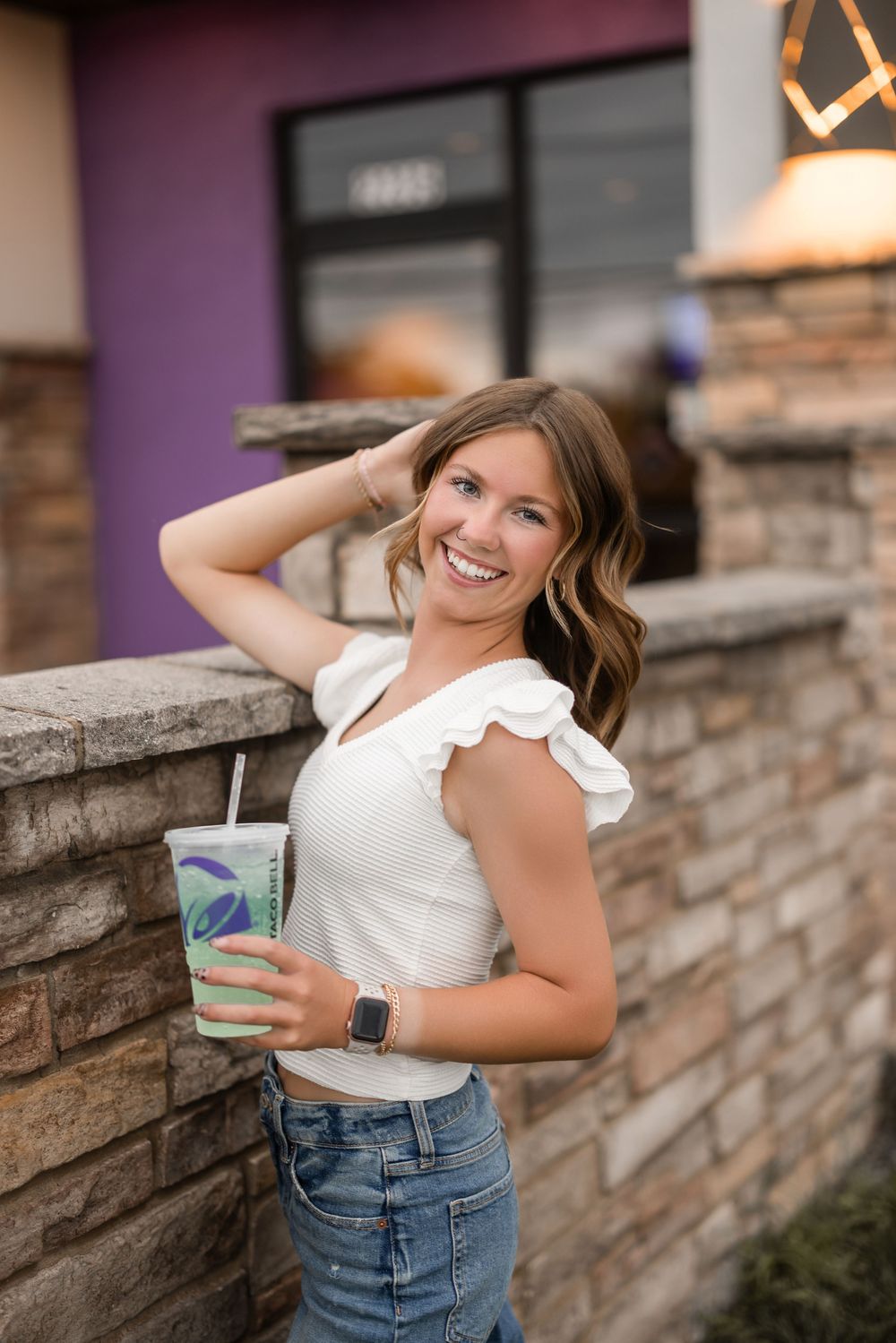 Person in white ruffled top and jeans poses with iced drink against stone wall while smiling outdoors.