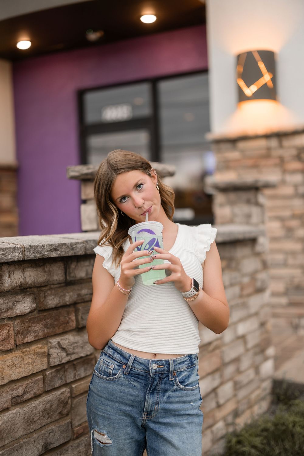 A person in a white ruffled top and distressed jeans drinks from a Taco Bell cup while standing by a stone wall.