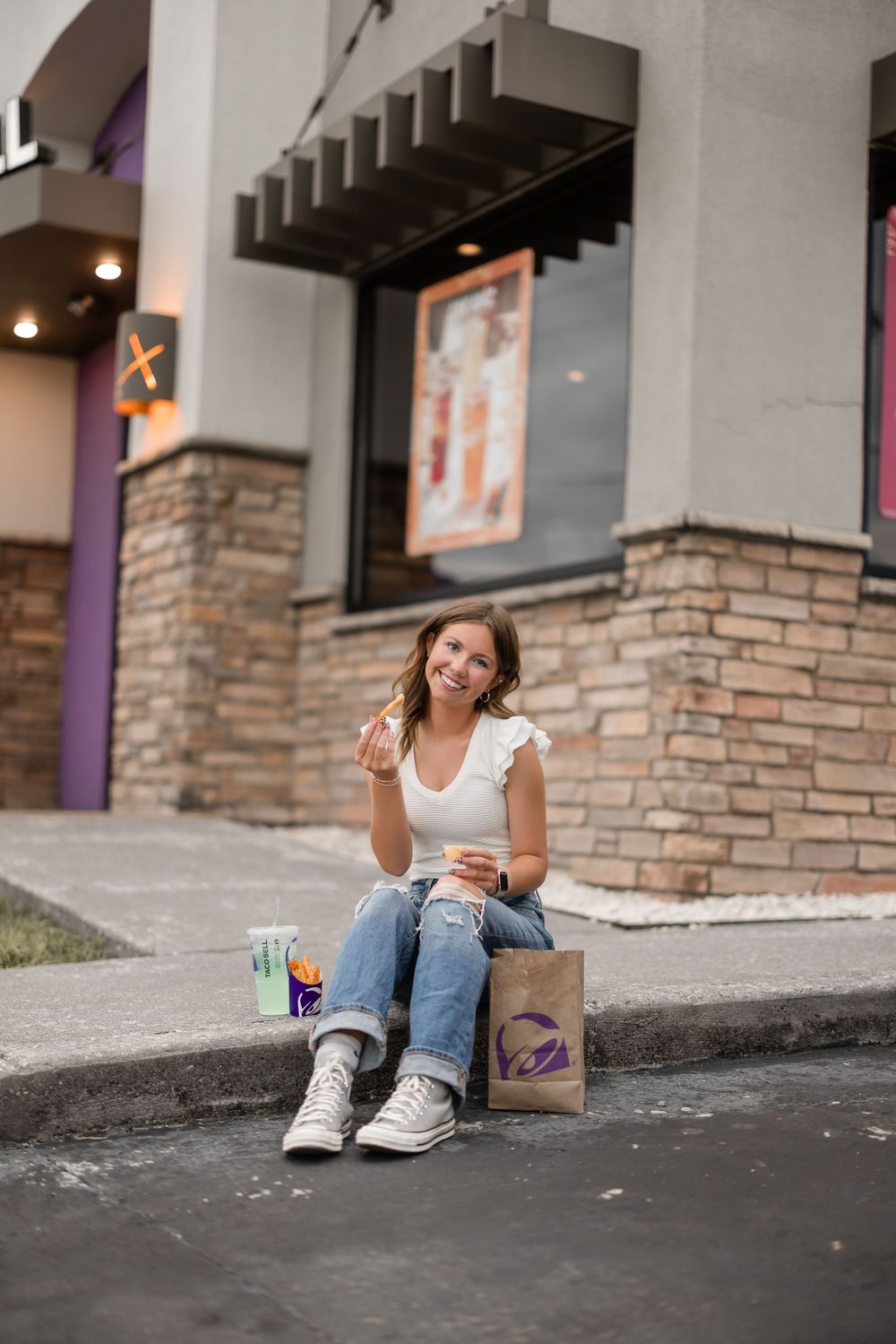 Someone sitting on a curb outside a brick building enjoying fast food and drinks.