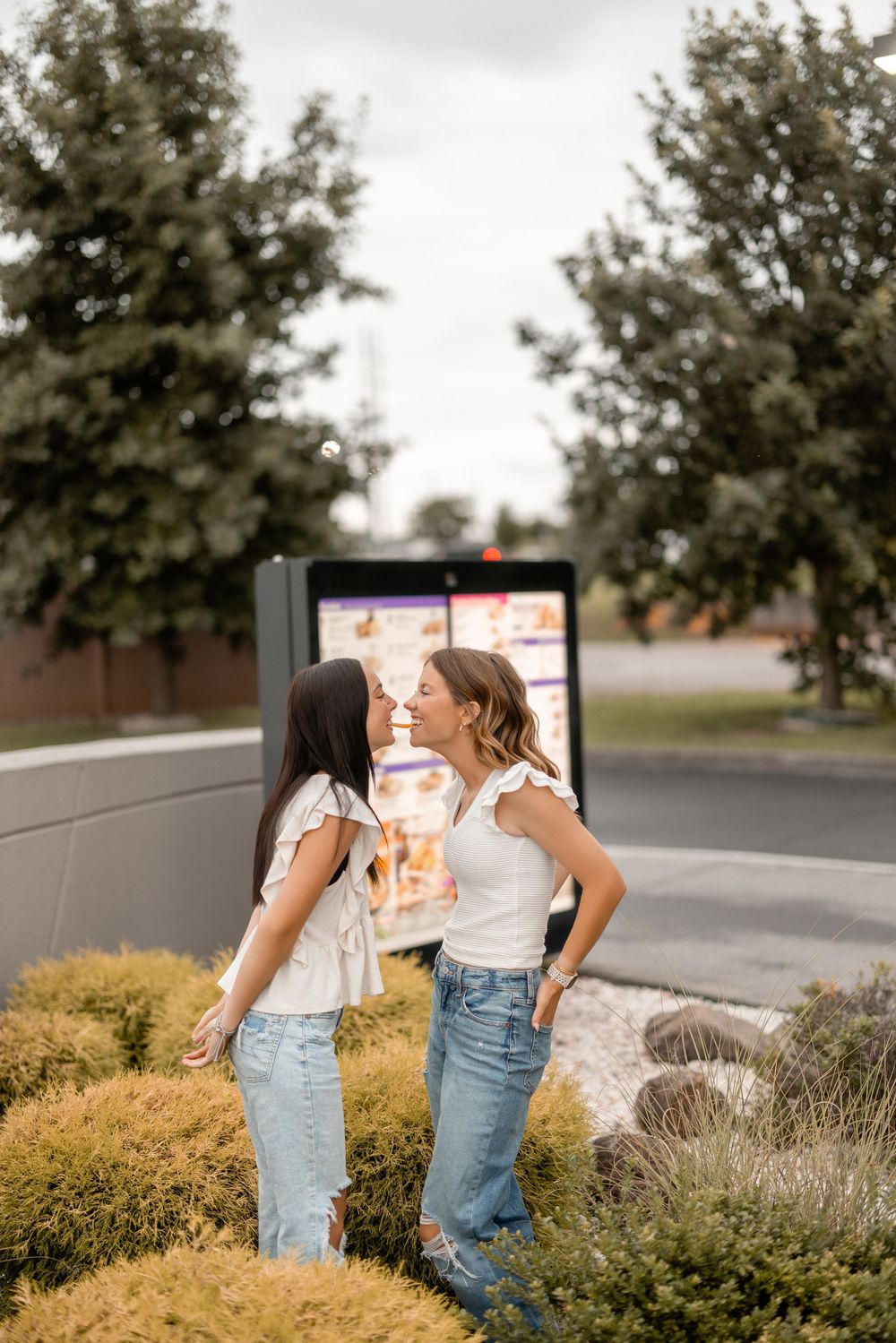 Two people in casual jeans and tops share a lighthearted moment together on a sidewalk next to decorative bushes.