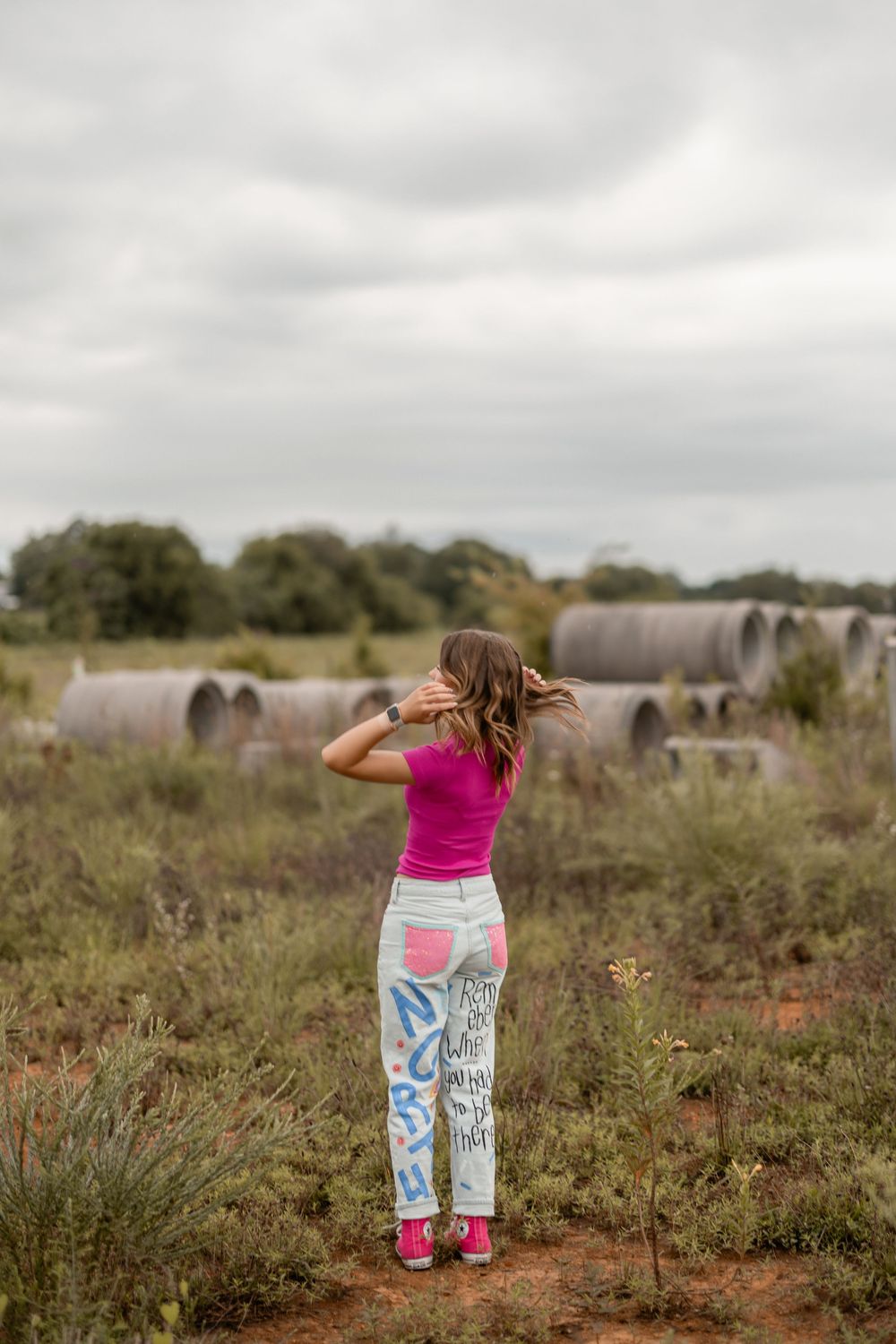 Woman in pink top and patterned pants gazes across an open field with large storage structures in the distance.