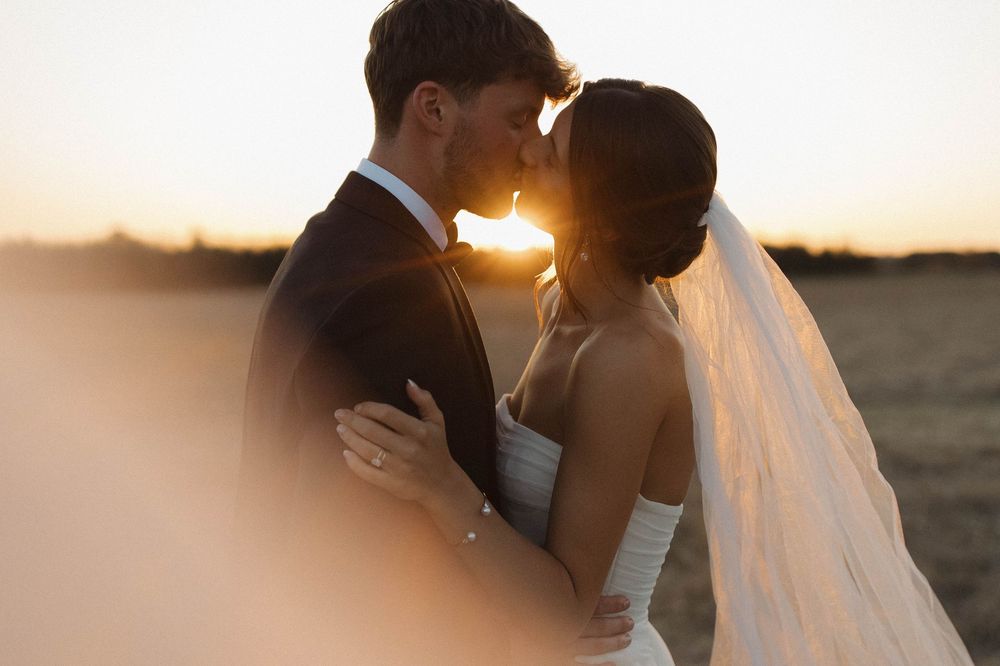Silhouetted newlyweds embrace during golden sunset with flowing veil creating romantic moment.