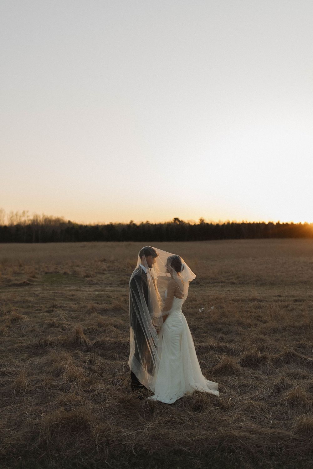 Bride and groom stand together in open field during sunset with long flowing veil.