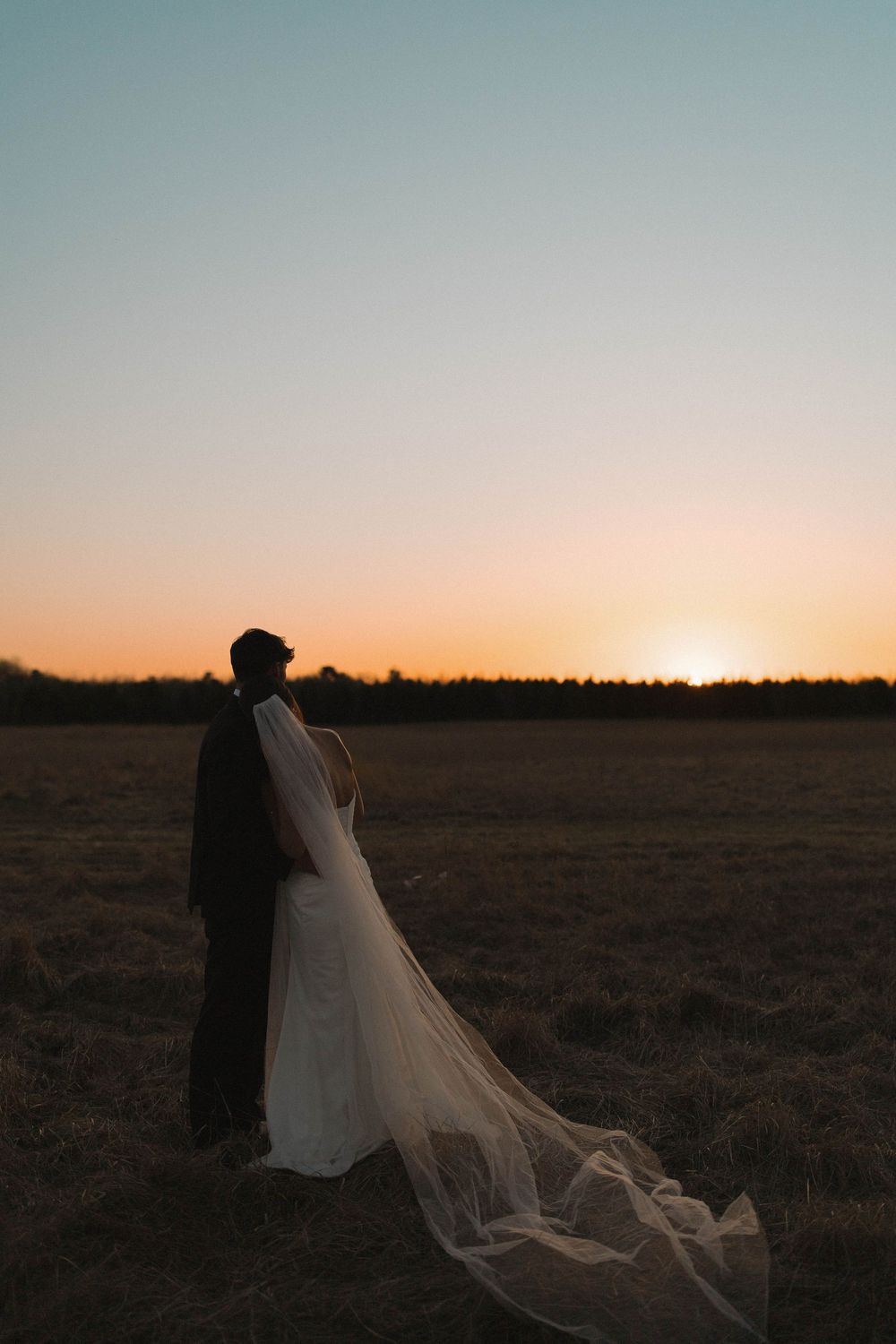 Wedding couple silhouetted against dramatic sunset with flowing cathedral veil in field.