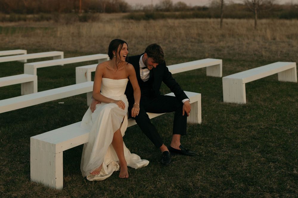 Couple sits together on white benches during outdoor wedding ceremony.