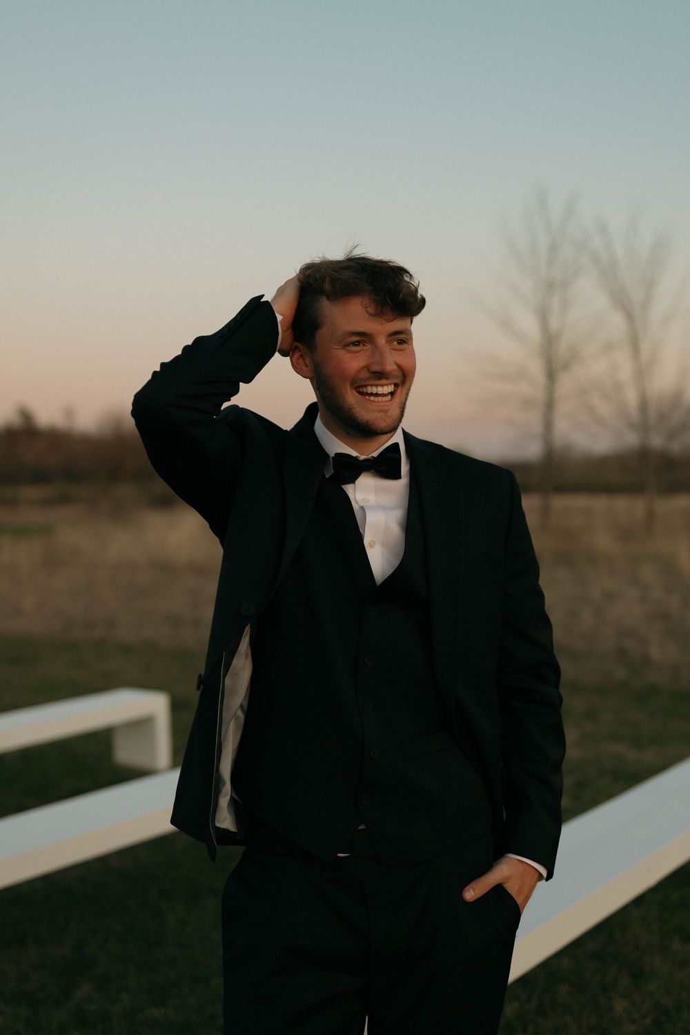 Groom in black tuxedo laughs while fixing hair during outdoor wedding portraits.