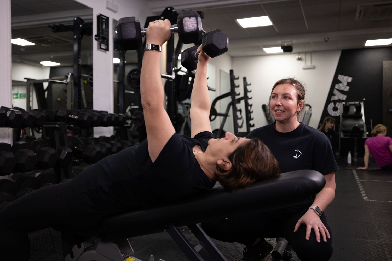 Personal trainer assists with weightlifting exercise on decline bench in modern fitness facility.
