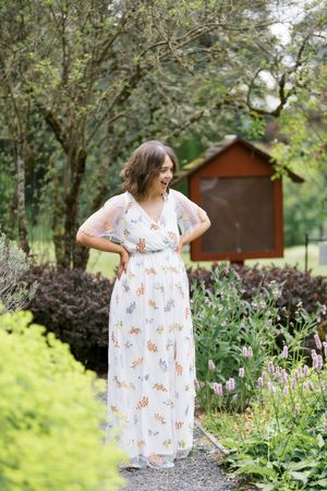 A woman in a white floral print dress poses in a garden setting with greenery and wooden structures in the background.