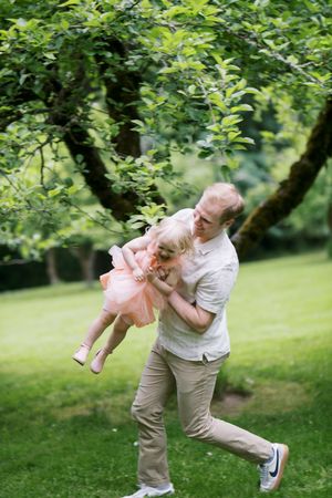 Playful moment captured in a lush garden as adult swings child wearing pink dress under green leafy trees.