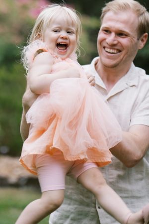 A playful parent twirling a small child wearing a peach-colored tulle dress in an outdoor garden setting.