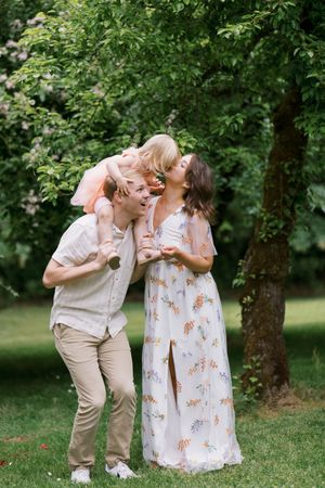 A touching family moment captured outdoors as three people interact playfully on a green lawn surrounded by leafy trees.