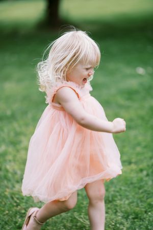 A small child in a peach-colored dress twirls playfully on green grass during a sunny day outdoors.