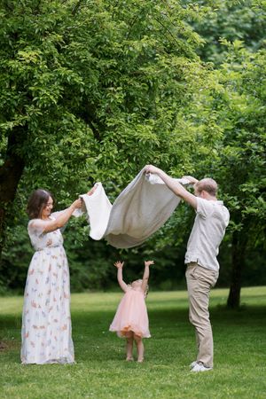 Family plays with blanket together in a lush green park on a summer day in matching white outfits.