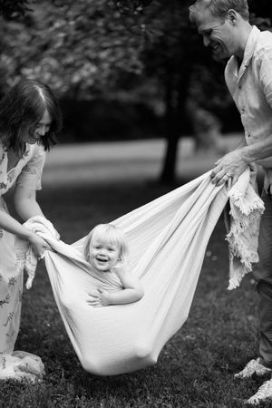 A joyful moment captured in black and white as a child swings happily in a makeshift hammock held by two adults outdoors.