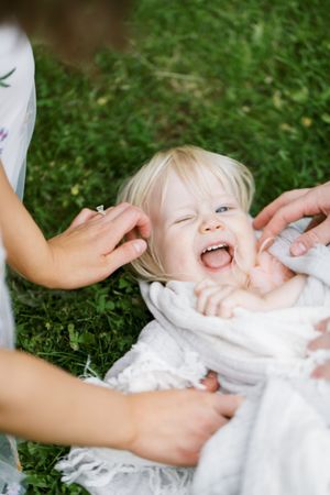 A laughing baby lying on green grass wearing white clothing while being gently touched by caring hands.
