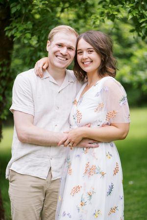 A couple poses together outdoors in casual spring attire against a green leafy background.