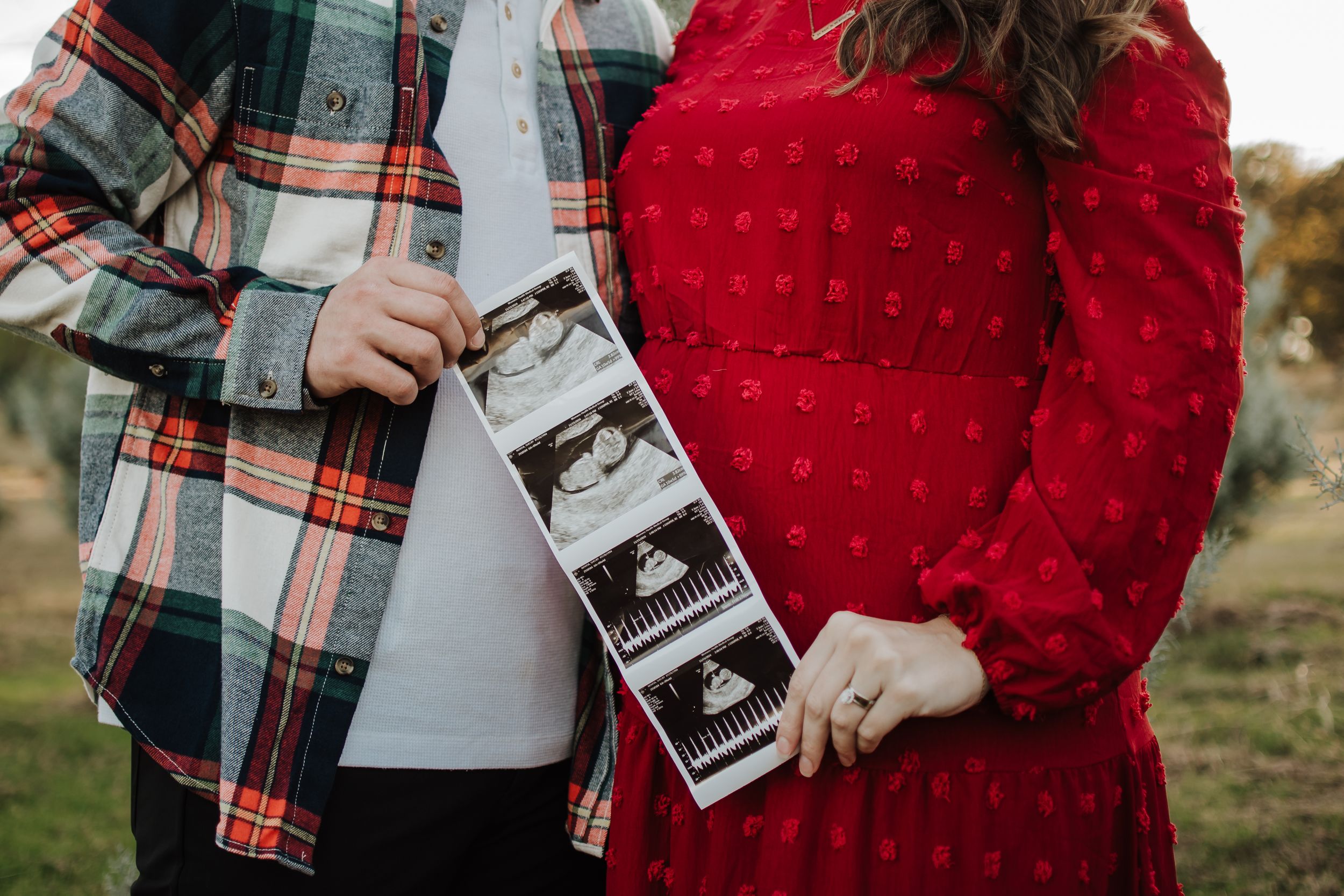 Close up of hands holding ultrasound photos with plaid shirt and red dress visible Tree Farm Mini Session Crowley Texas