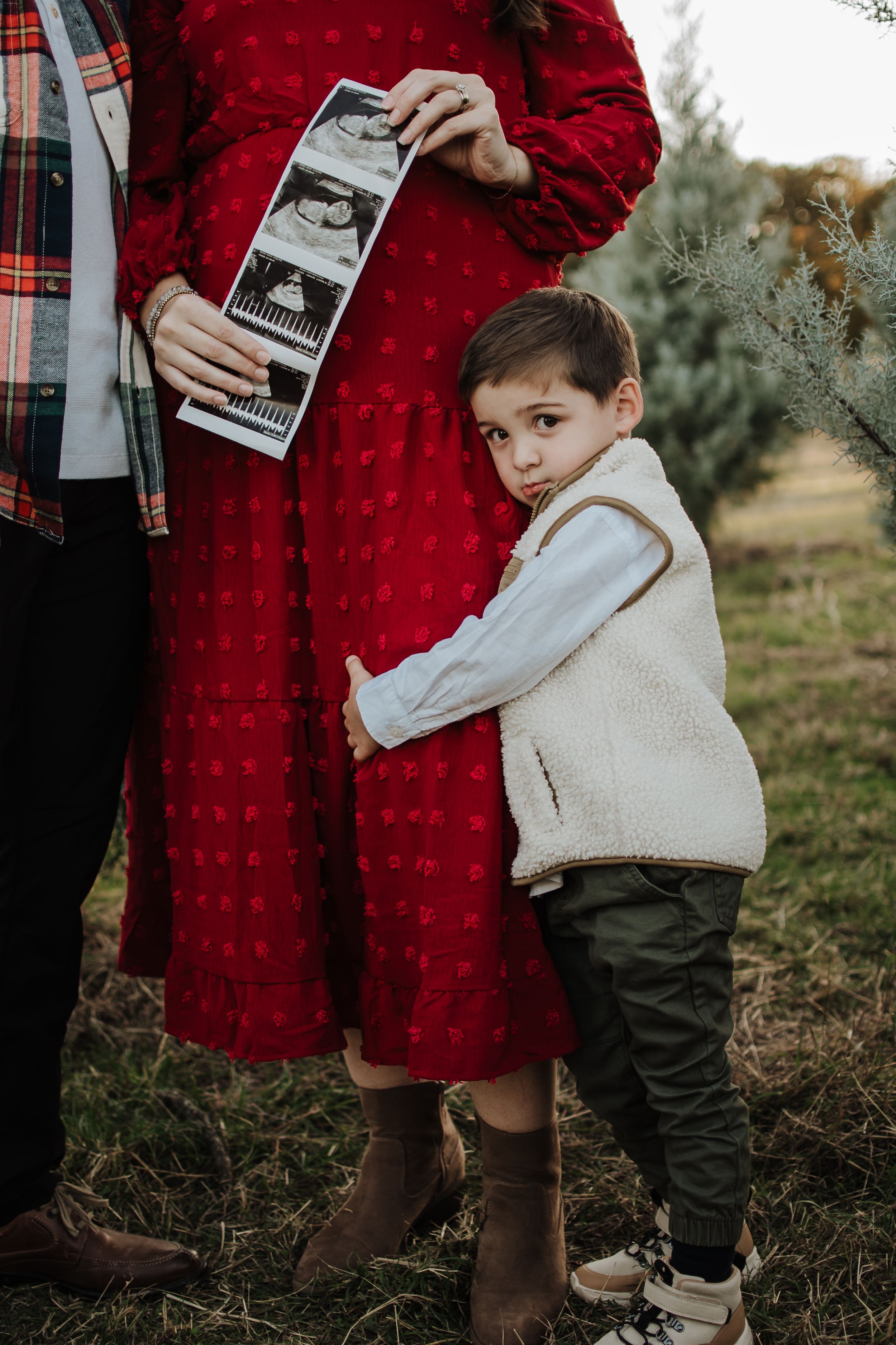 Child in cream vest hugging person in red dress holding ultrasound photos outdoors. Tree Farm Mini Session Crowley Texas