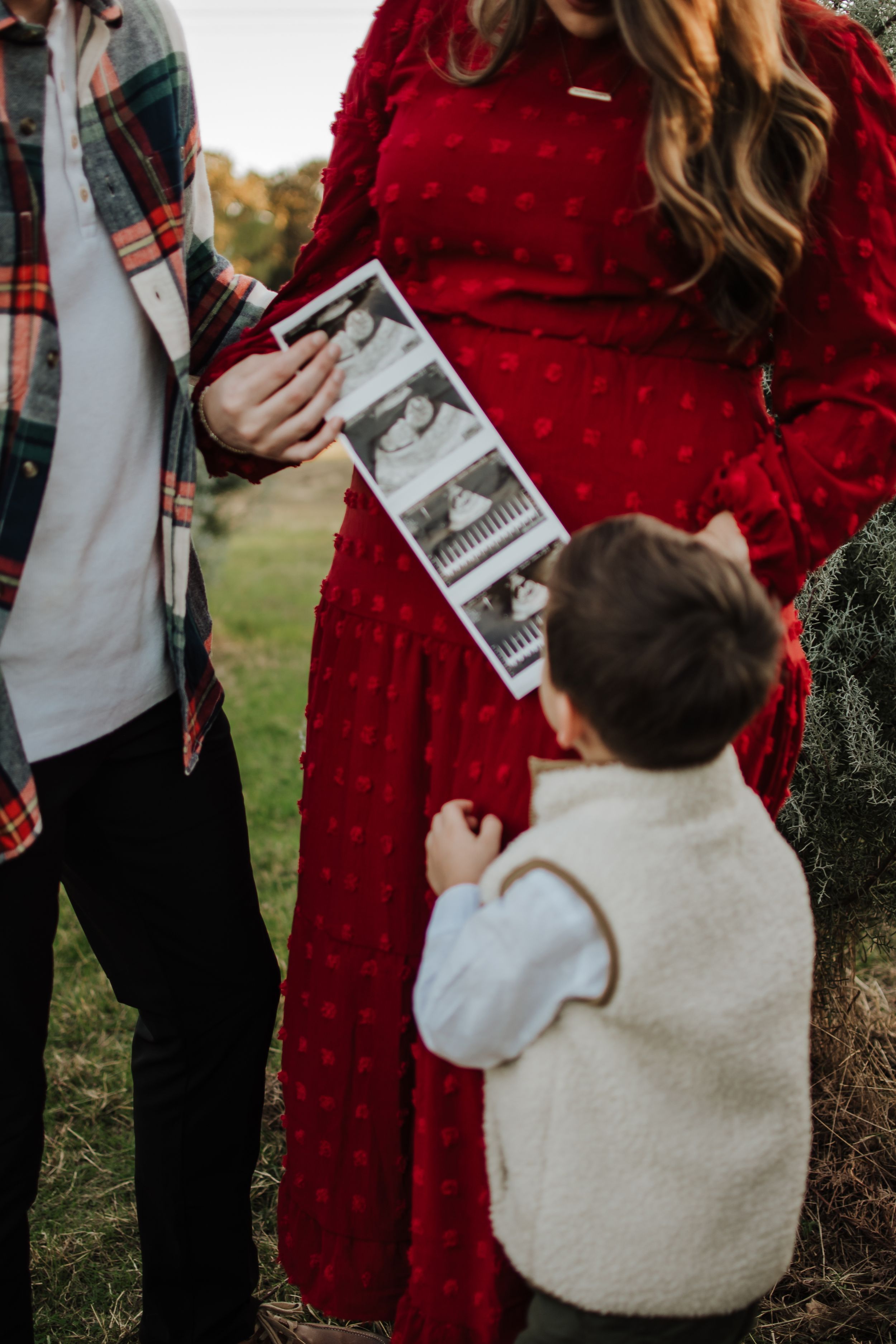Family looking at ultrasound strip while child in cream vest holds onto red dress. Tree Farm Mini Session Crowley Texas
