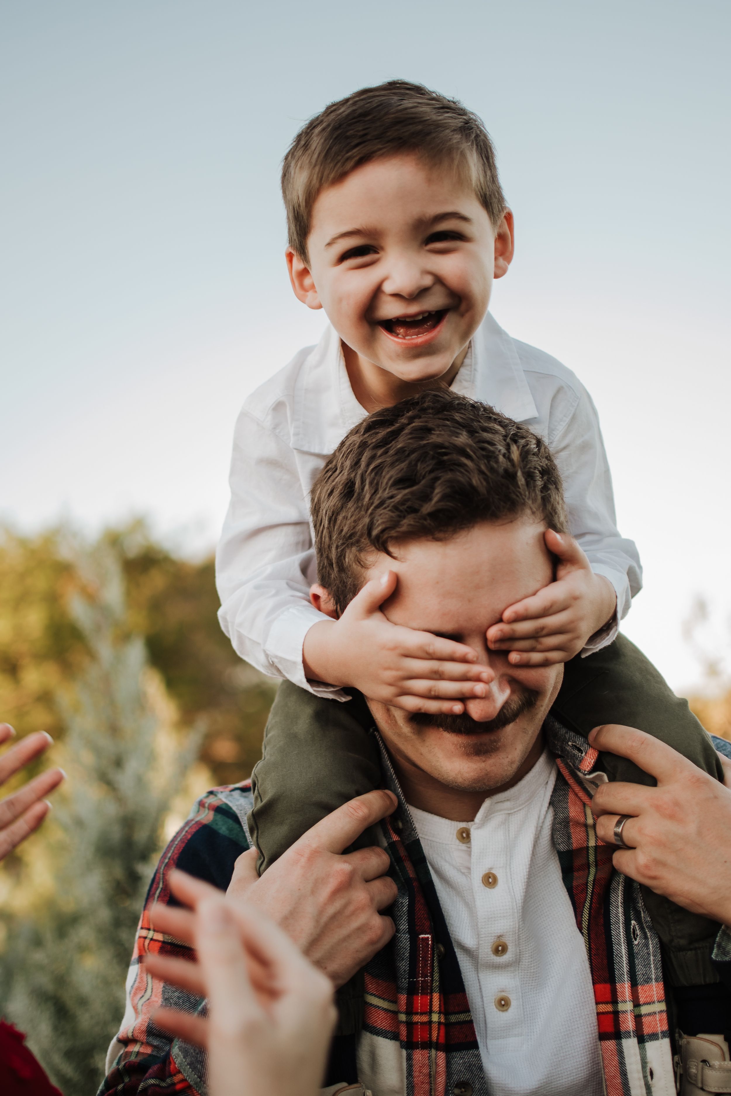 A playful moment of laughter and joy as a child covers someone's eyes while riding on shoulders outdoors.