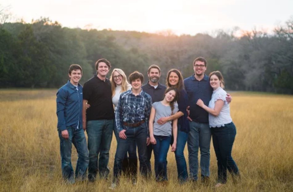 Group of people wearing casual attire standing together in a sunlit grassy field at sunset.