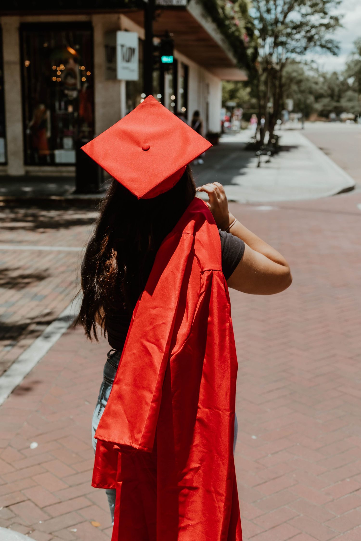 Graduate carrying red cap and gown walks down brick sidewalk on sunny day.
