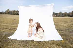 Two subjects in light clothing sit by a large white backdrop sheet set up in an open grassy field on a sunny day.
