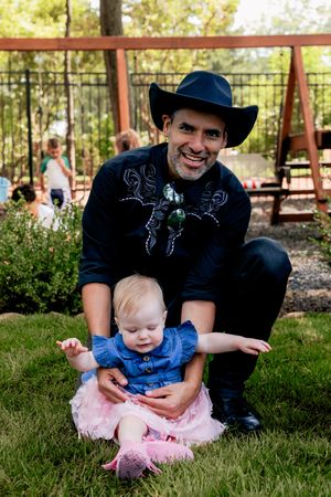 A playful outdoor scene with a baby in a tutu and denim dress on grass, sitting with someone wearing black.