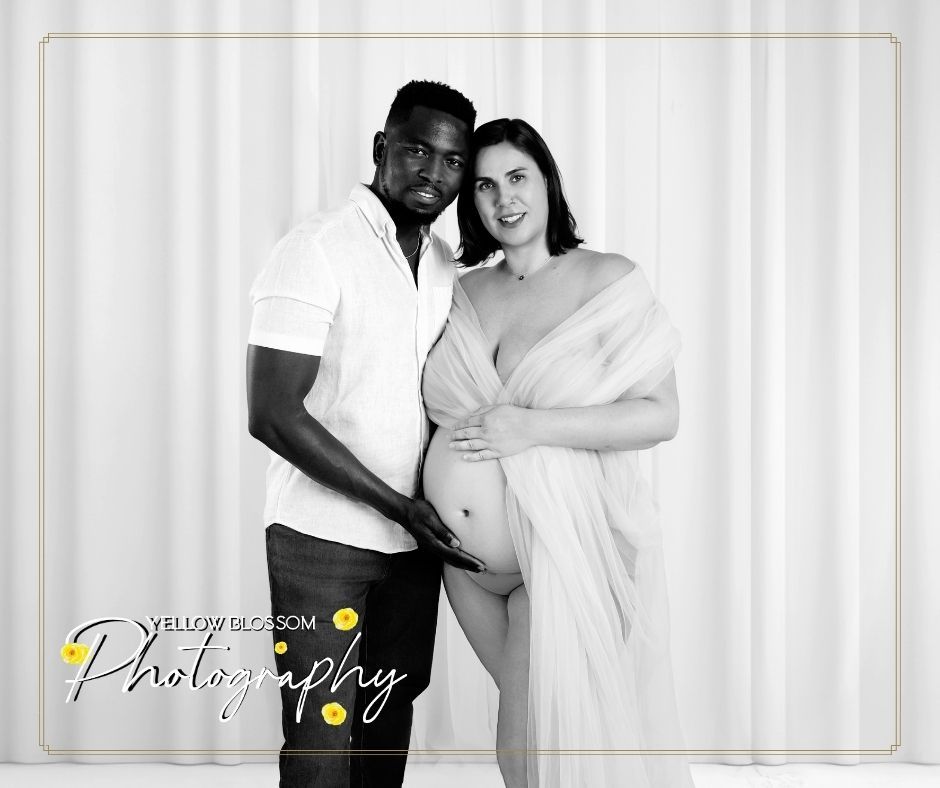 A black and white maternity photo of a couple standing together against a white curtain background.