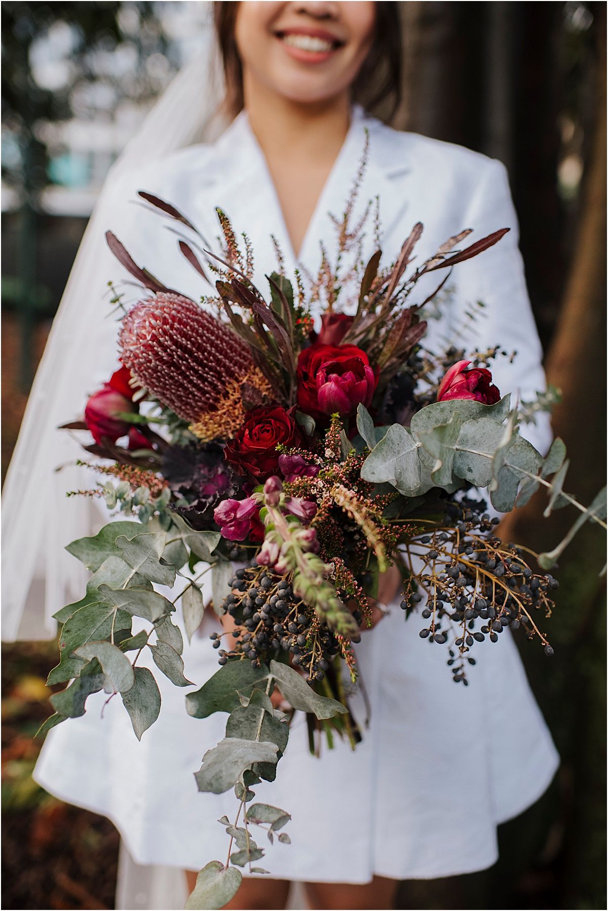 A stunning bridal bouquet featuring deep red roses, protea, dried grasses and cascading eucalyptus against white fabric.