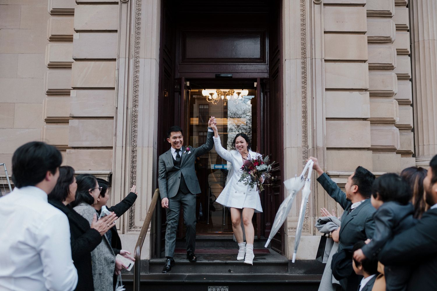 Happy couple exits building with guests tossing confetti on the steps during wedding celebration.