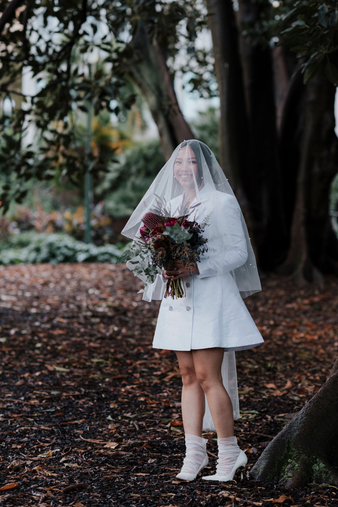 A person in a short white wedding dress with boots and veil holds a deep red bouquet while standing on fallen leaves.