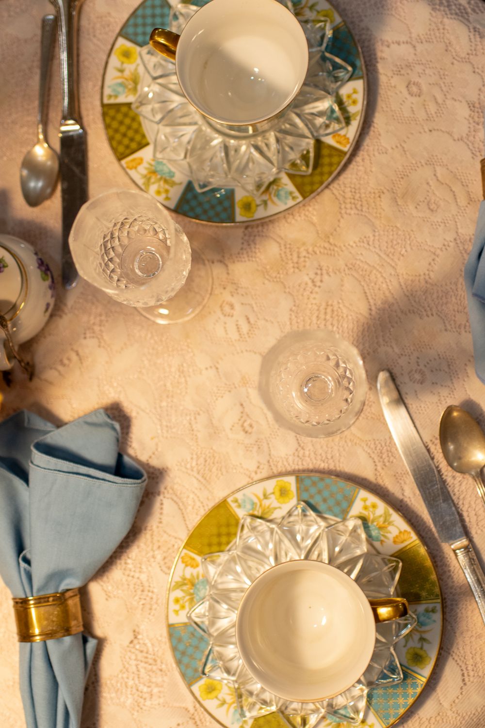 Elegant tea table setting with crystal cups on colorful floral plates, silver spoons, and blue napkins on white lace cloth.