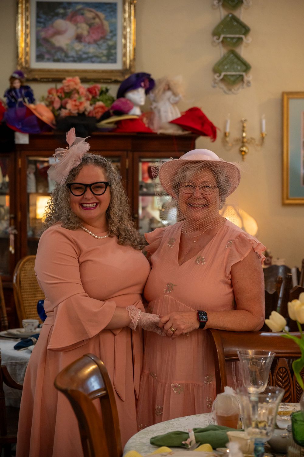 Two women in matching pink dresses and hats smile together at an elegant tea party setting.