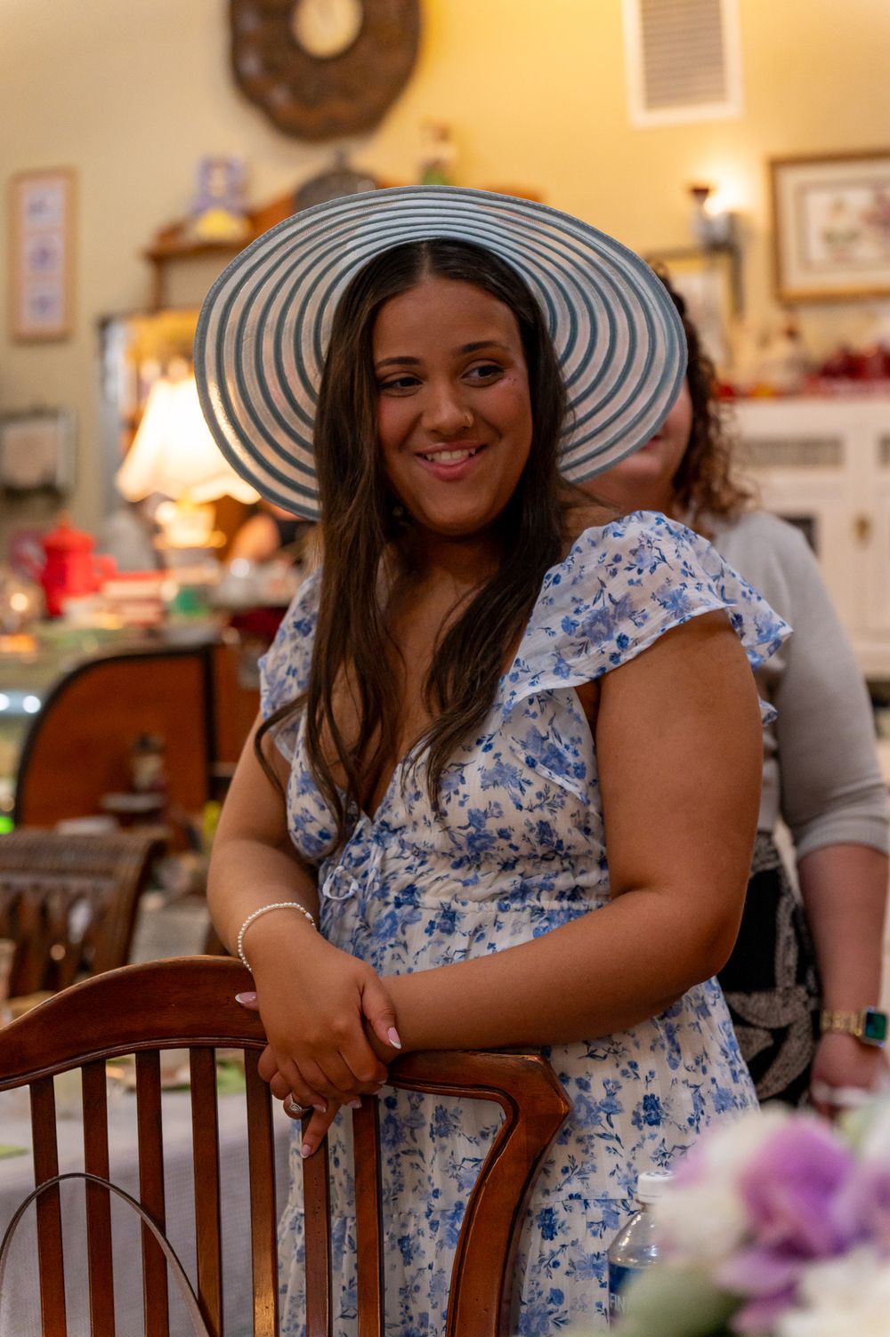 Smiling woman in blue floral dress and wide-brim striped hat posing by a wooden chair in a cozy decorated room.