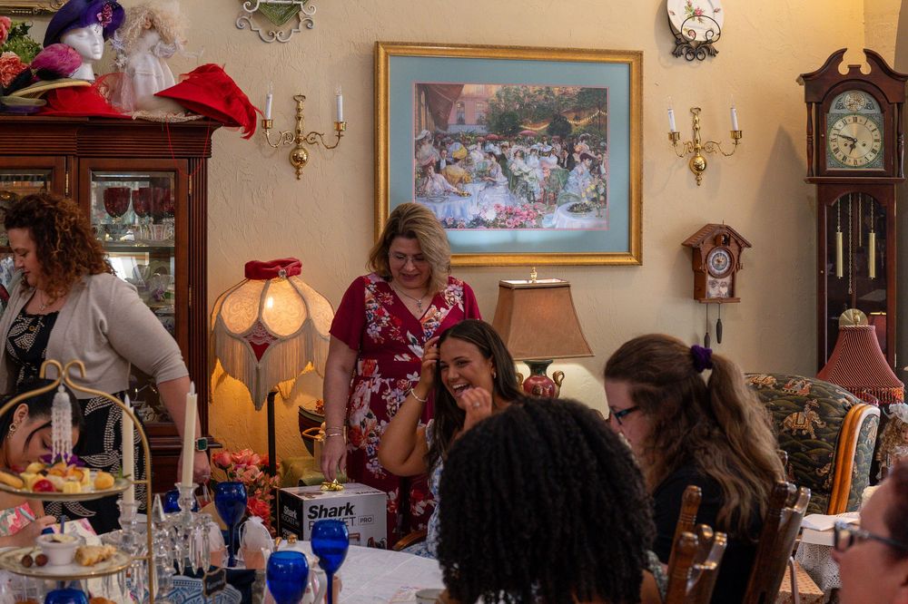 Women gather around a decorated table at an elegant tea party with vintage decor, paintings, and a grandfather clock.
