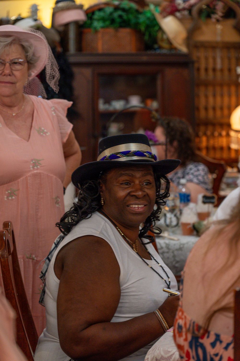 Woman smiling in a decorated hat at an elegant tea party gathering with guests in vintage attire.