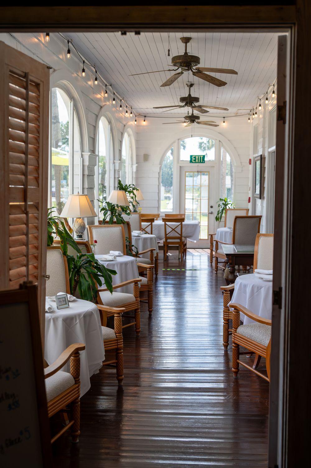 Elegant restaurant hallway with arched windows, ceiling fans, white tablecloths and wooden floors lit by string lights.