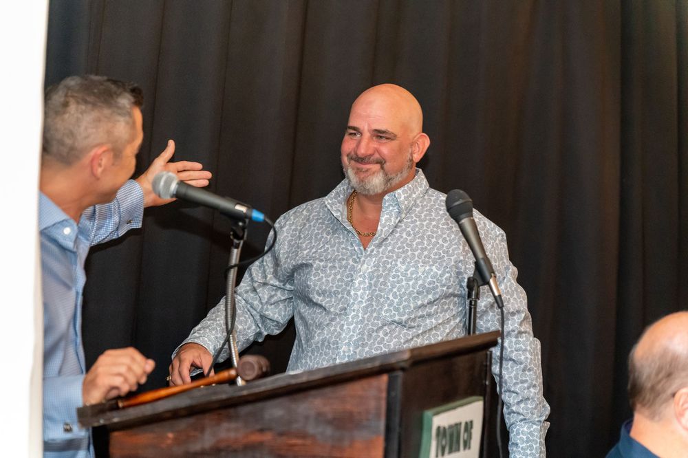 Speaker at podium wearing checked shirt smiles and gestures during presentation on stage with dark curtain background.
