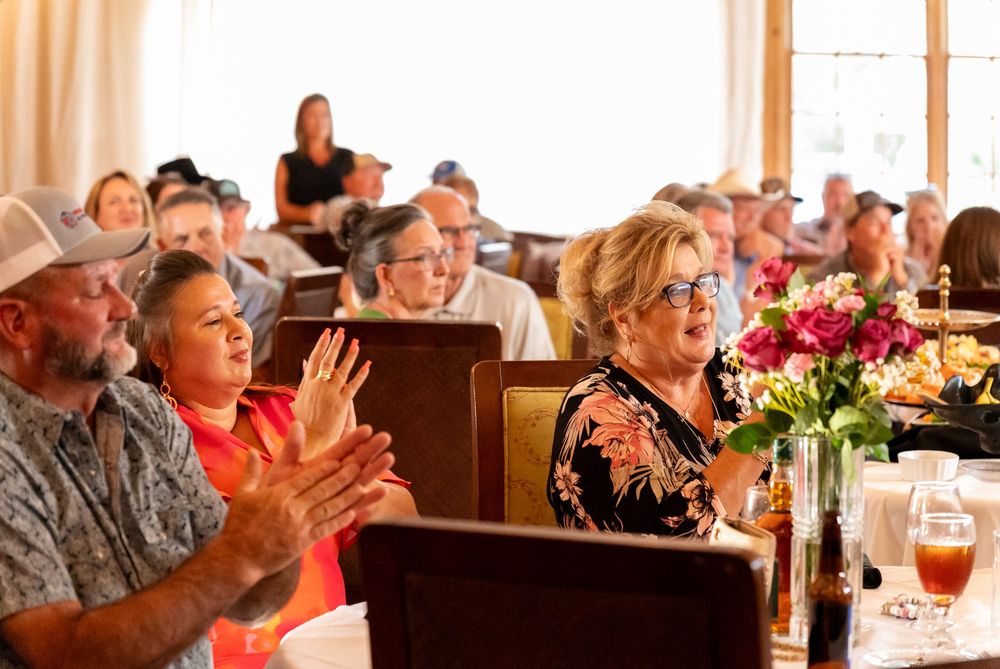 Guests applaud during a dinner event with floral centerpieces in an upscale restaurant setting.
