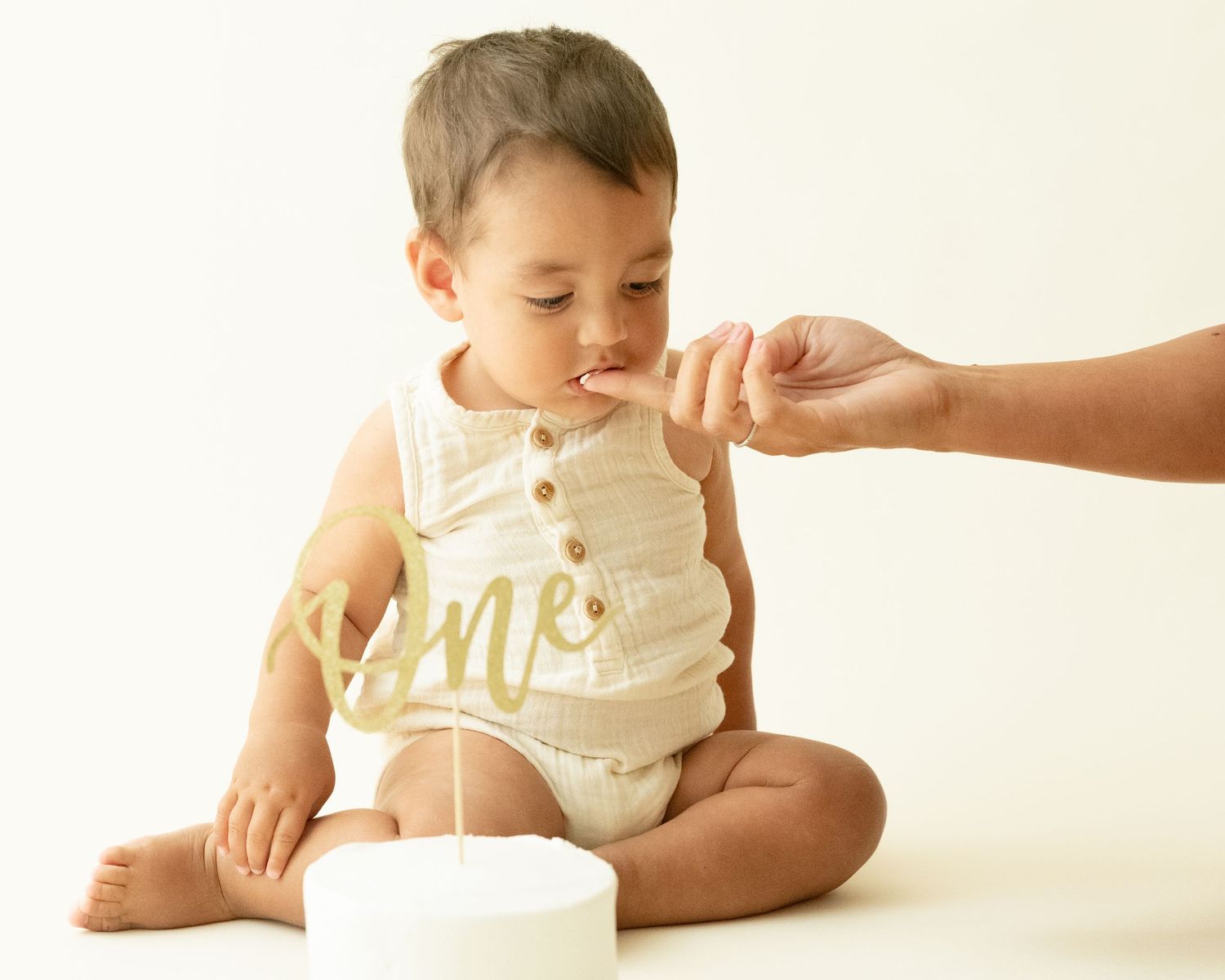 Small child in cream-colored romper sits on white surface while adult hand offers food.