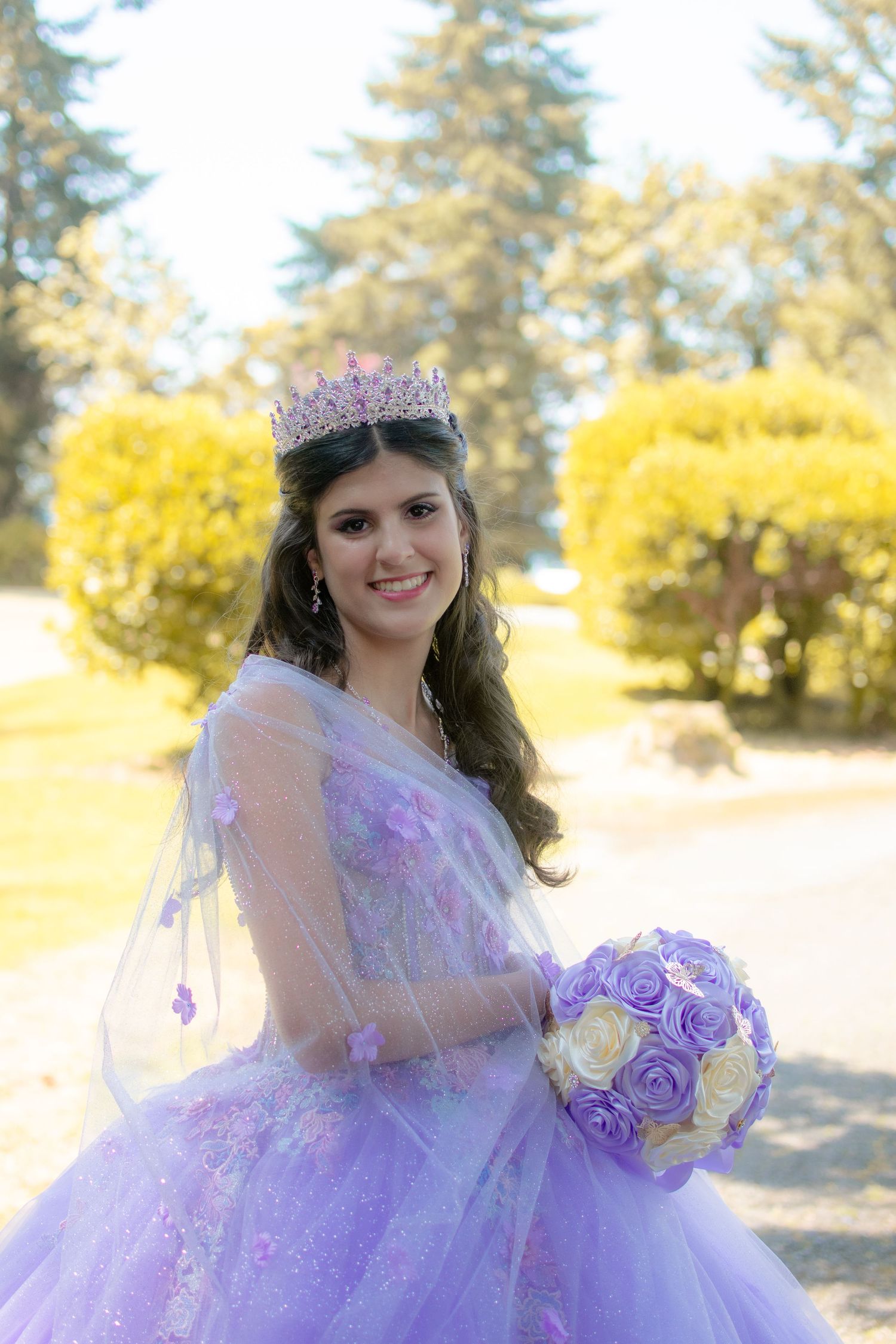 Person in lavender ball gown with crown holding flower bouquet in sunny garden.