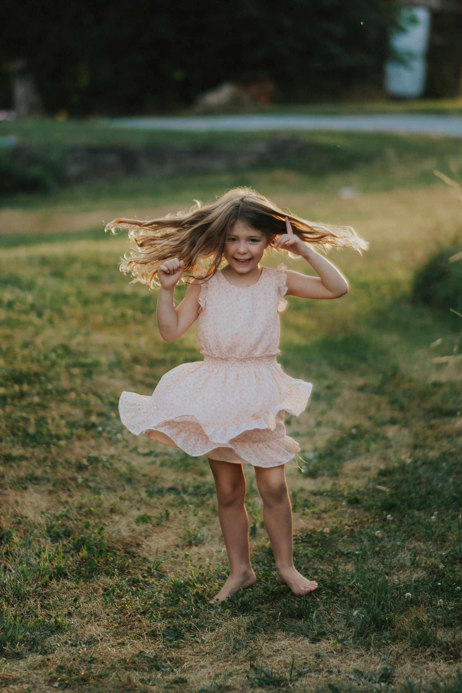 A small child in a pink dress twirling and dancing joyfully on grassy ground during a summer day.