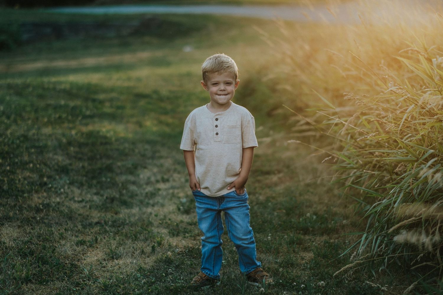 A young child stands in jeans and a beige shirt against golden sunlit grass during sunset.