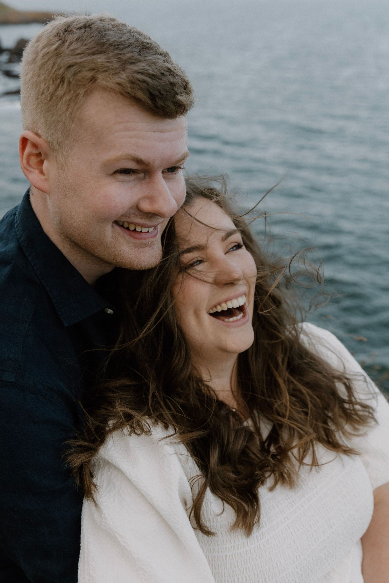 Happy couple laughing together against ocean backdrop during sunset photoshoot.