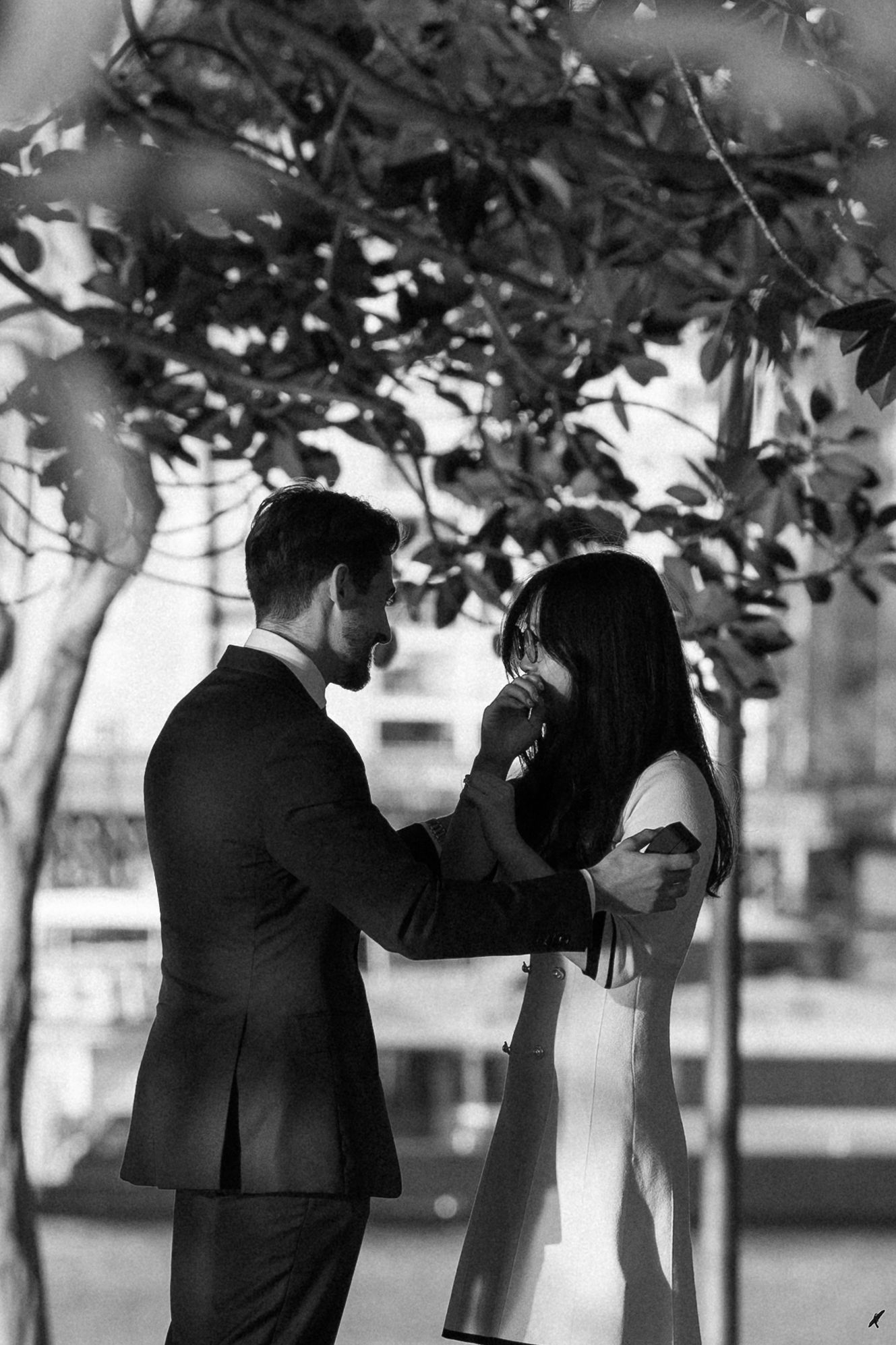 A black and white photo shows people in formal attire standing beneath trees in an outdoor setting.
