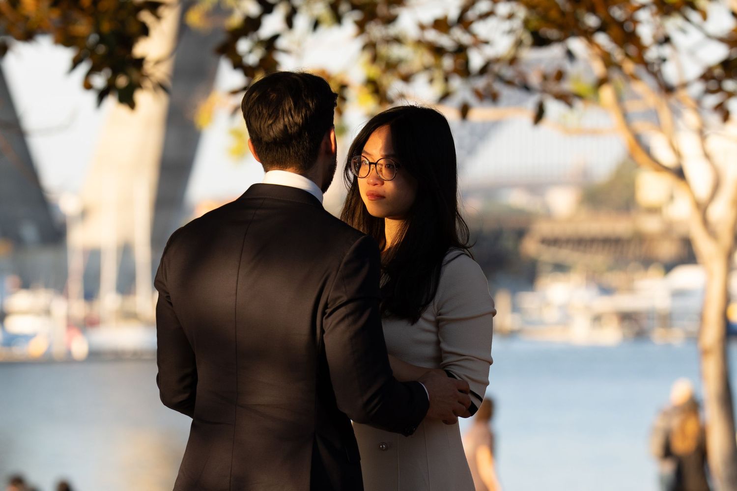 A couple embraces during a golden hour photoshoot by the waterfront with autumn trees in the foreground.
