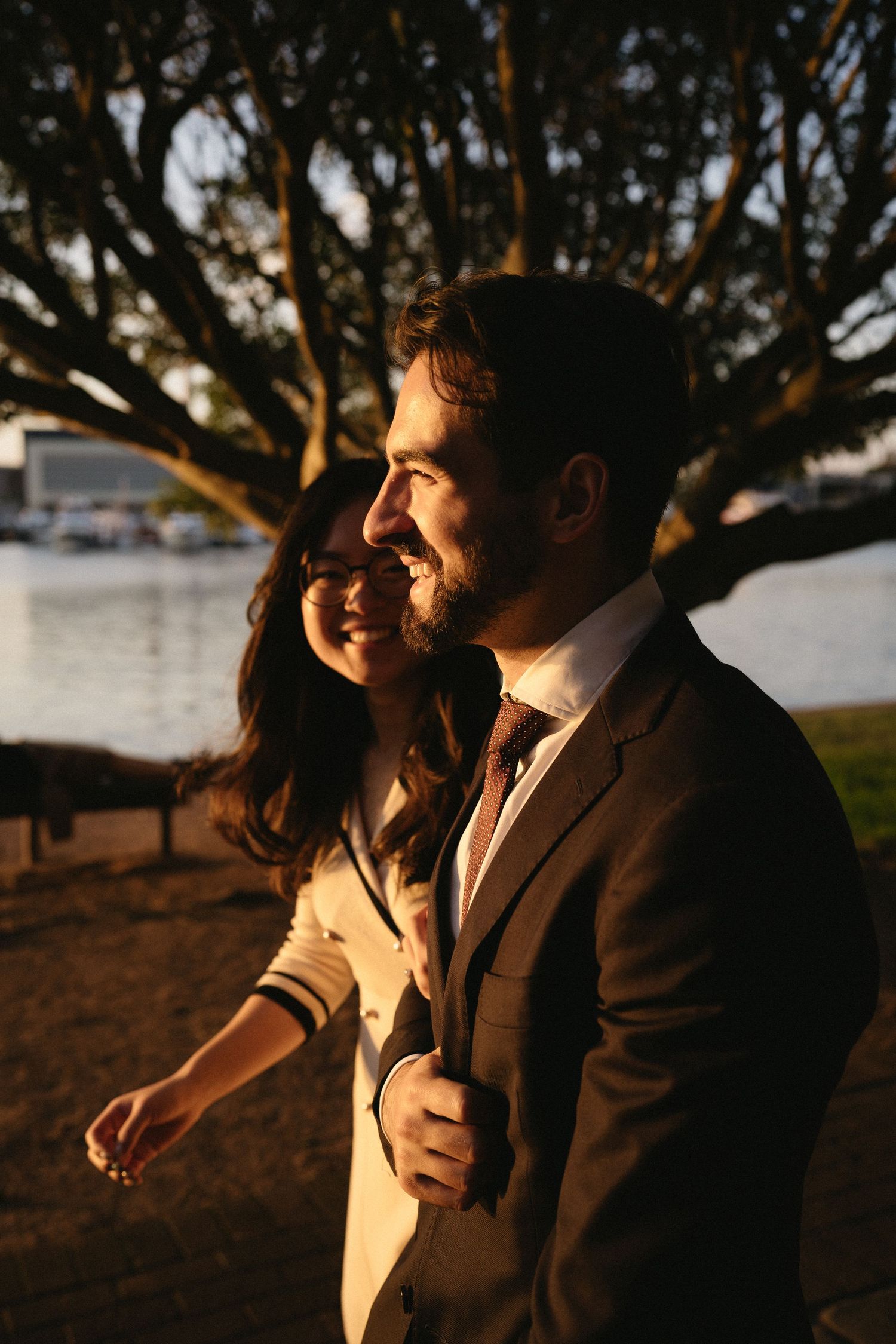 A couple sits together outdoors at sunset, sharing an intimate moment beneath trees along the waterfront.