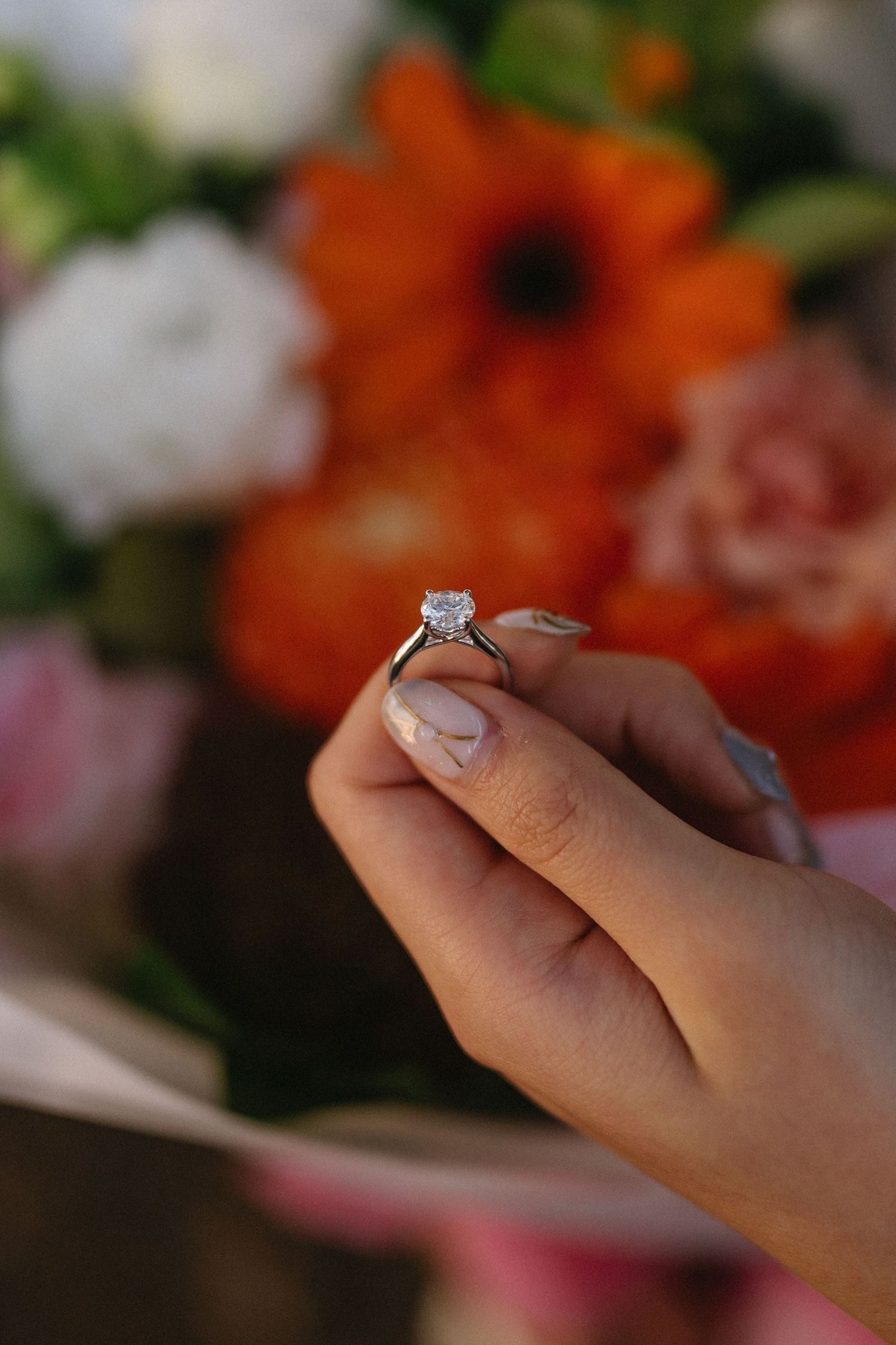 Hands hold a sparkling diamond engagement ring against a blurred background of orange and pink flowers.